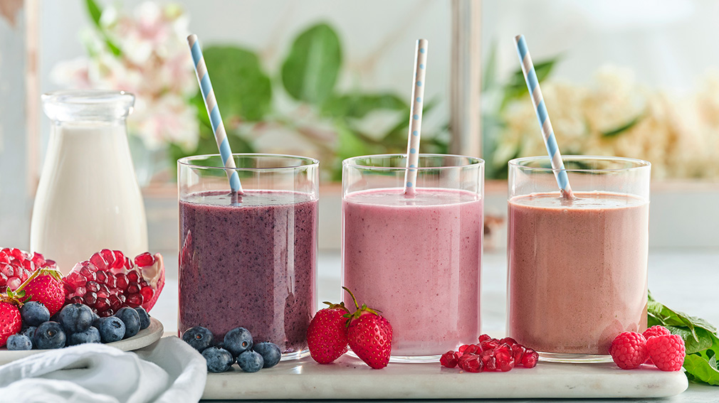 Three glass tumblers are filled with Milk and Pomegranate Delight, Creamy Raspberry Swirl and Banana Blueberry Kefir smoothies, with a blue and white paper straw in each.
