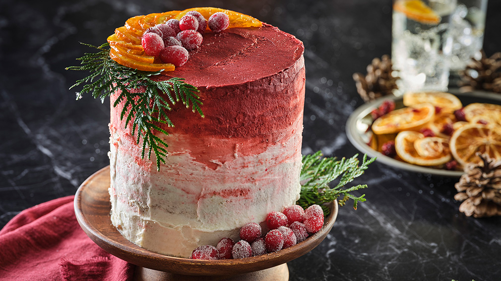 A tall Vanilla Sour Cream Cake with red and white Buttercream Frosting sits on a wooden cake stand, decorated with sprigs of pine and sugared cranberries.