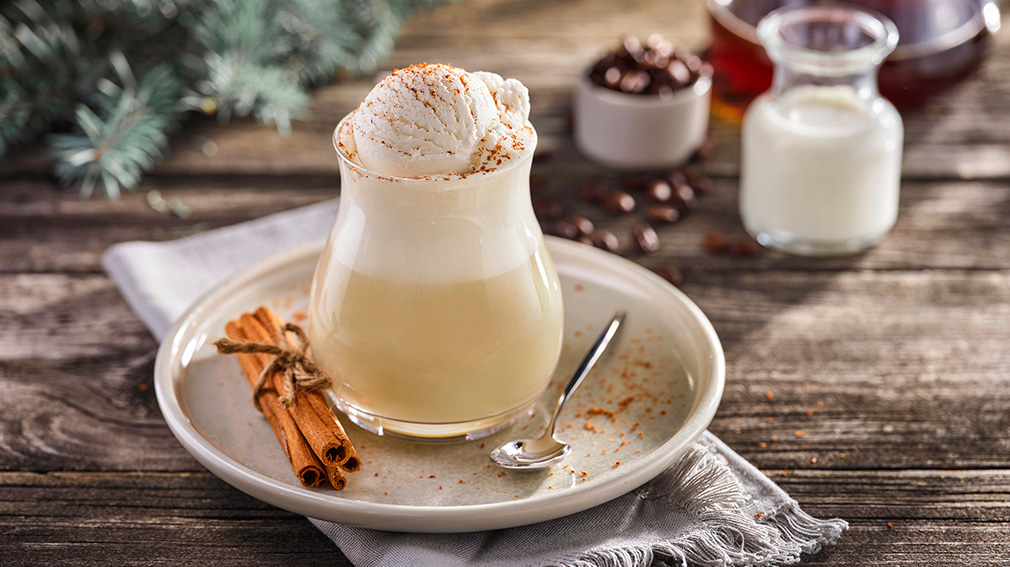 A glass with Creamy Holiday Coffee Nog, with a tied bunch of cinnamon and a silver sits on a beige earthenware plate on a grey napkin on a wooden table.