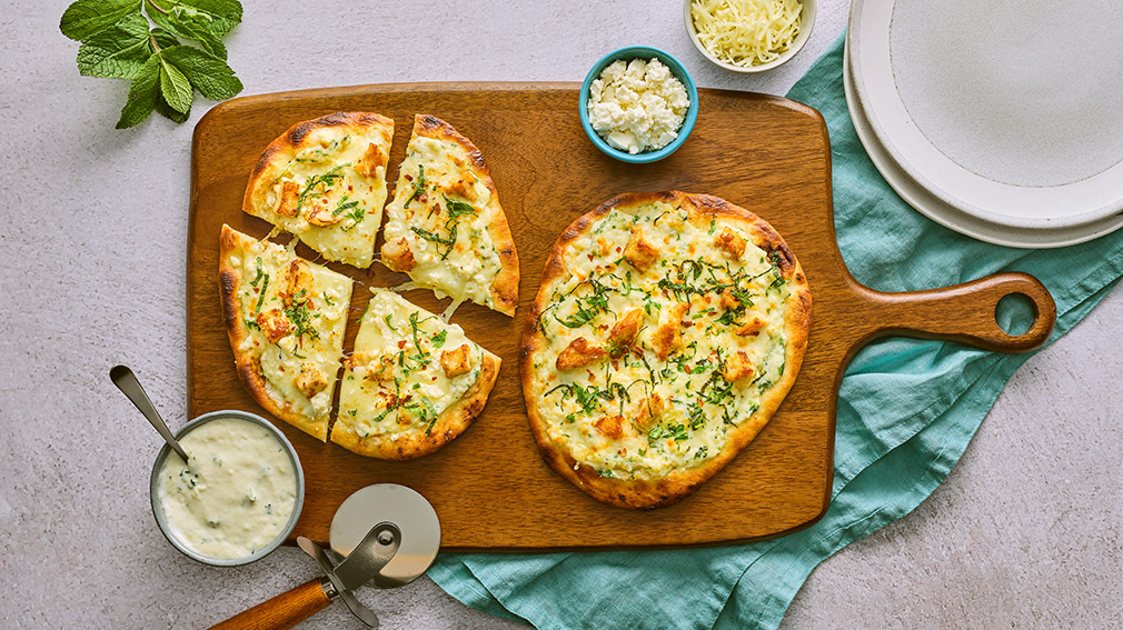 An overhead view of two small naan pizzas on a wooden serving board on top of a blue napkin surrounded by small round bowls of cheese, garlic cream sauce, plates, a pizza cutter, and a spring of mint.