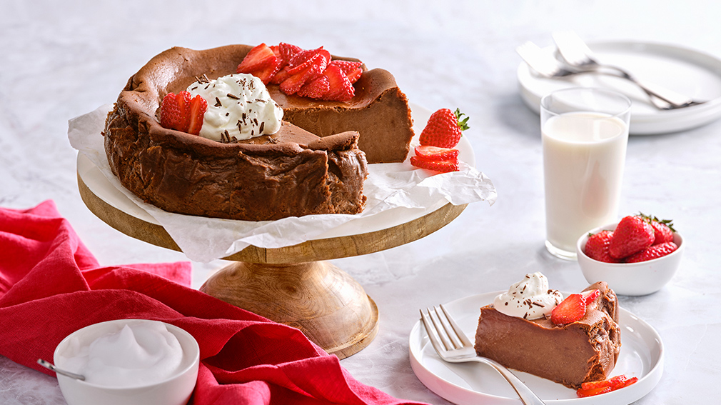 A chocolate basque cheesecake topped with whipped cream and fresh strawberries resting on parchment paper on top of a light wooden cake stand, a red napkin and a small white bowl of whipped cream.