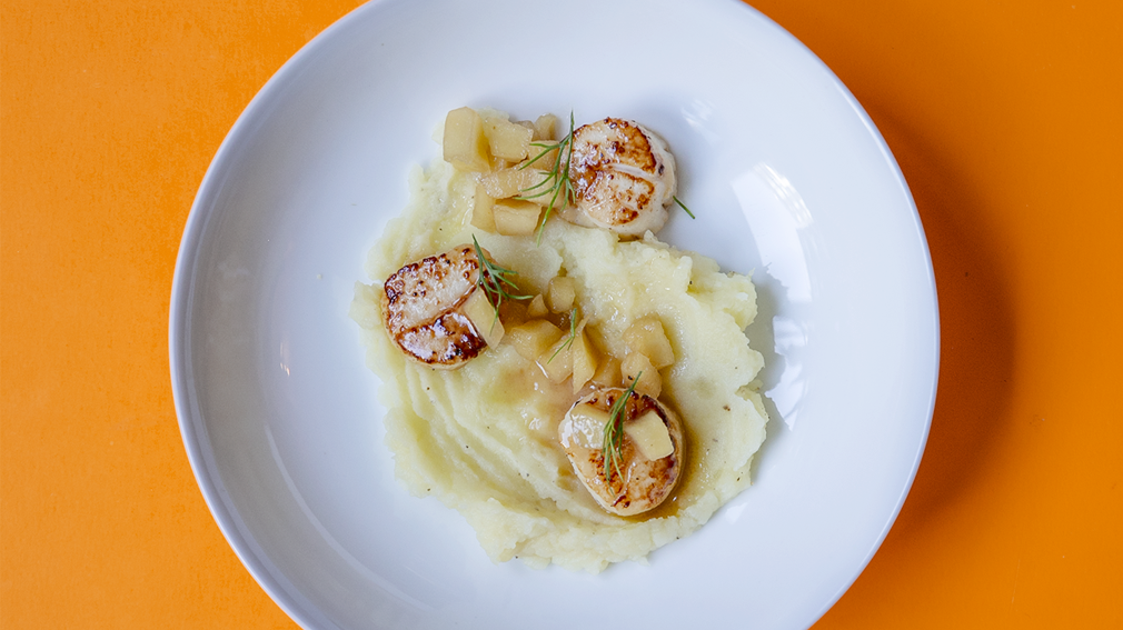 Three scallops sitting in a bowl over mashed potatoes and a golden apple sauce.