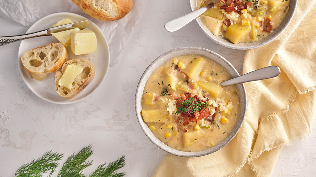 Two small grey earthenware bowls with spoons are filled Bacon & Corn Chowder with Cheddar and sit on a yellow napkin with a small plate with sliced baguette and butter.