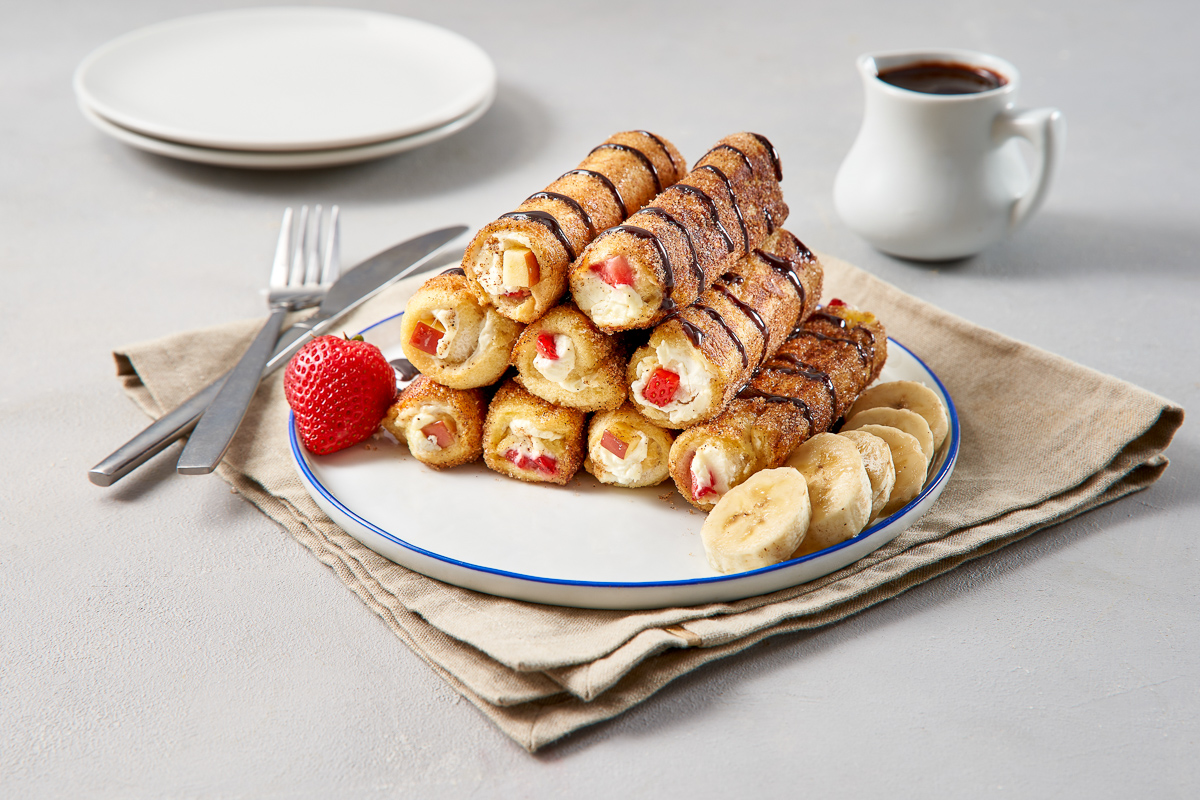 A white plate with nine Buttermilk French Toast Roll ups stacked in a pyramid formation. One whole strawberry and six banana slices also on the plate.