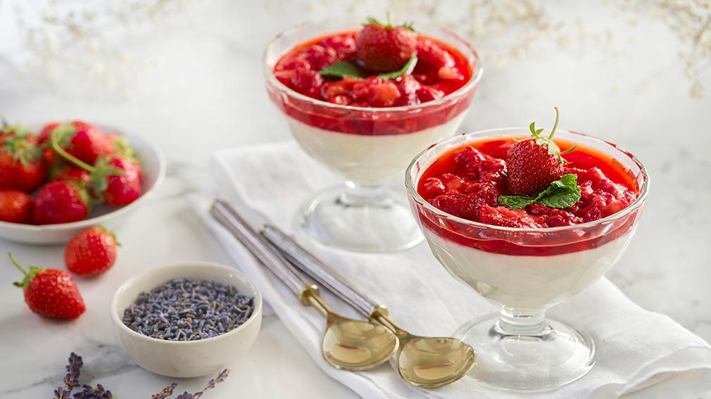 Two footed small dessert bowls are filled with Creamy Strawberry Panna Cotta and sit on folded white tablecloth next to two gold and silver spoons.