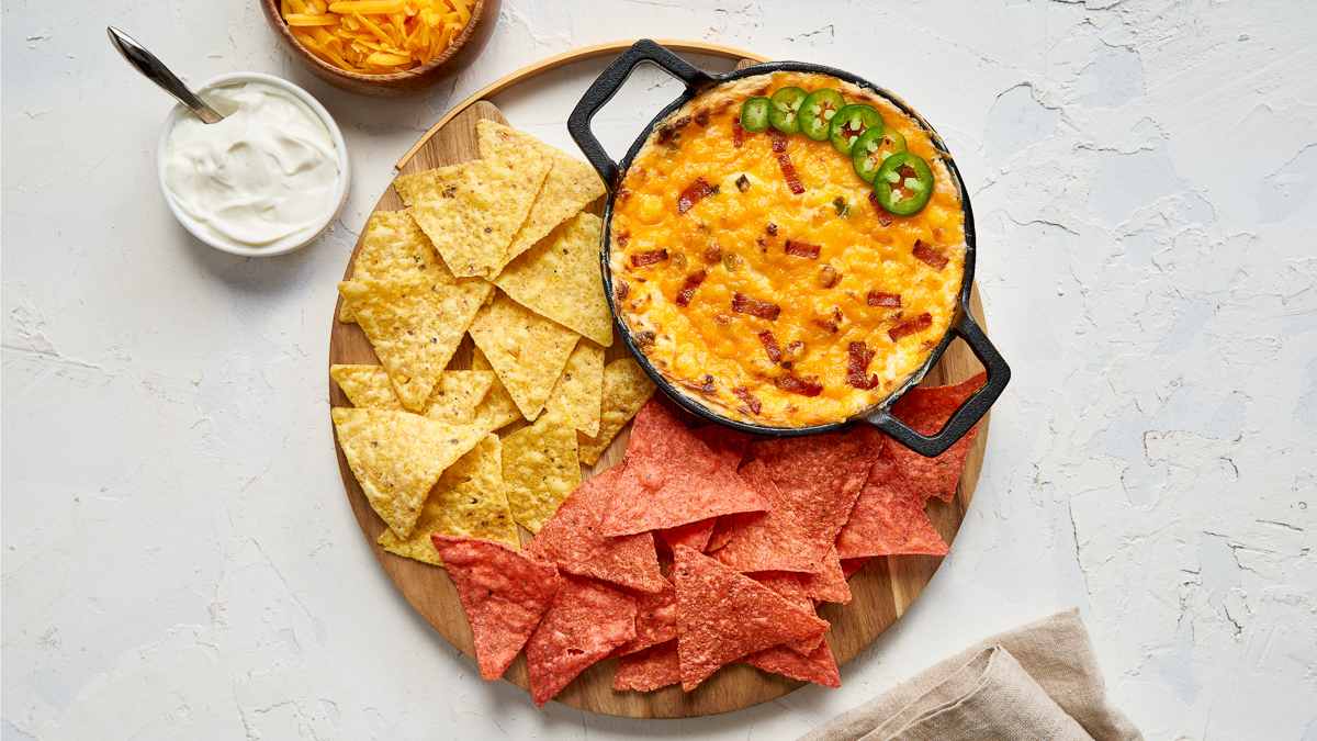 Black bowl with Jalapeno Popper Dip, topped with six jalapeno slices and a sprinkle of bacon bits. The bowl is on a plate which is filled with red and yellow tortilla chips for dipping.