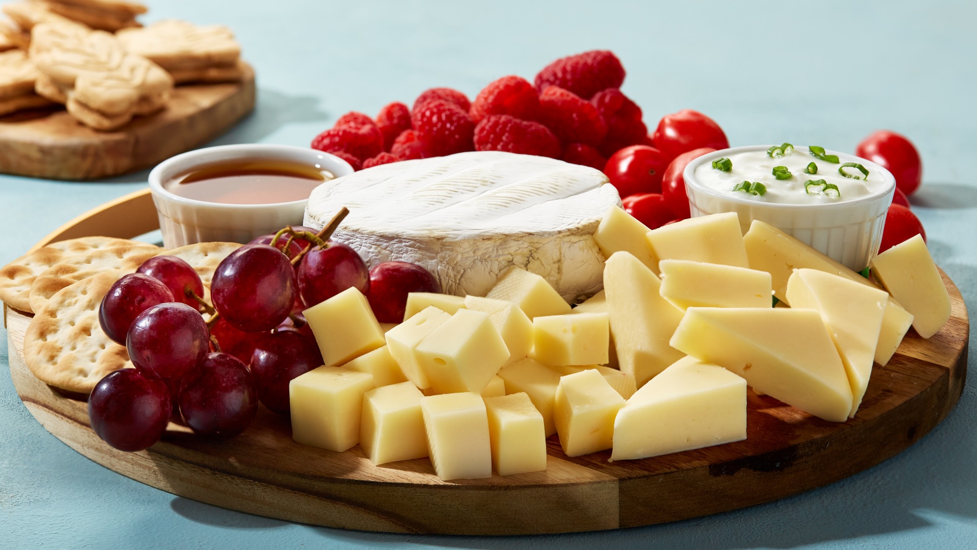 Round board with a brie wheel in the middle, surrounded by cut cheese, grapes, sour cream dip, fruit and crackers
