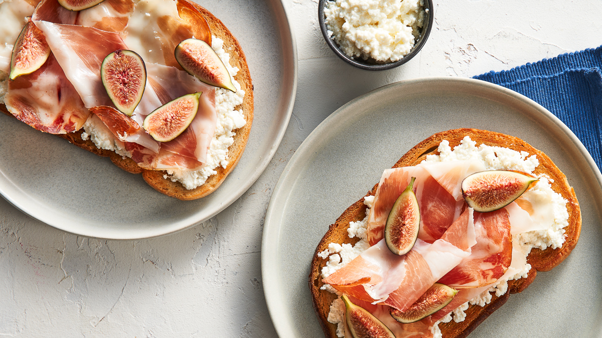 Two white plates, each with a slice of rye bread topped with a layer of ricotta cheese, a layer of prosciutto, a few wedges of fig and a drizzle of honey.