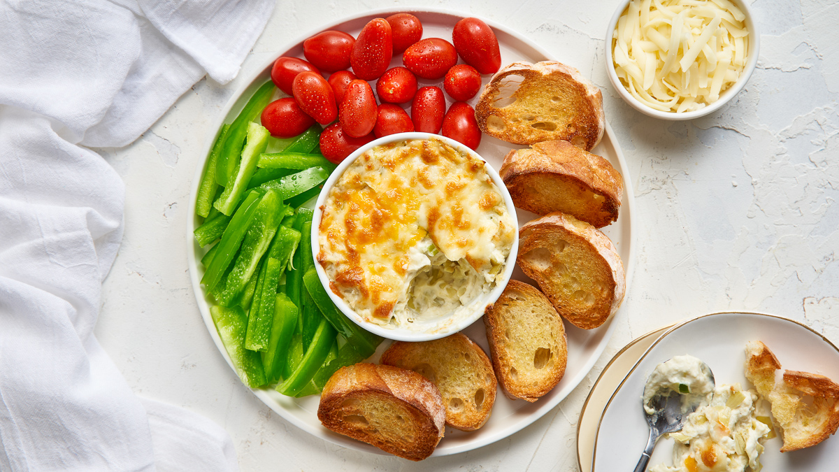 White bowl filled with three cheese artichoke dip in the centre of a white plate. The bowl is surrounded by green peppers, cherry tomatoes and baguette slices.