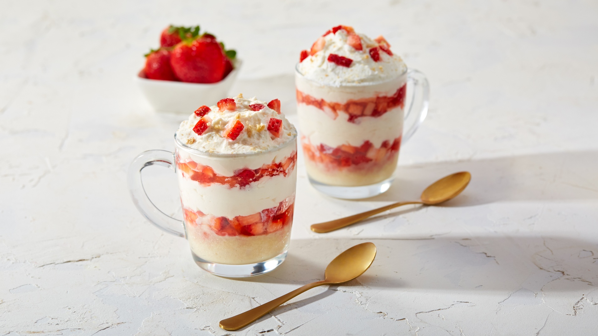 2 clear glasses with layers of crushed graham crackers, strawberries and whipped cream