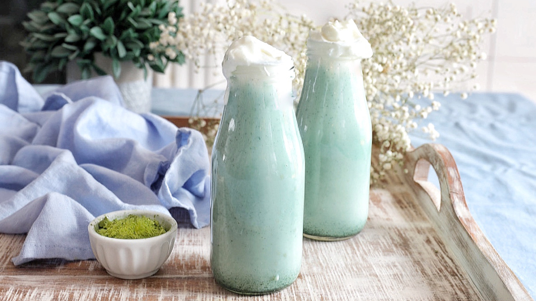 Two milk bottles with green/blue smoothie with whipped cream on top on serving tray.