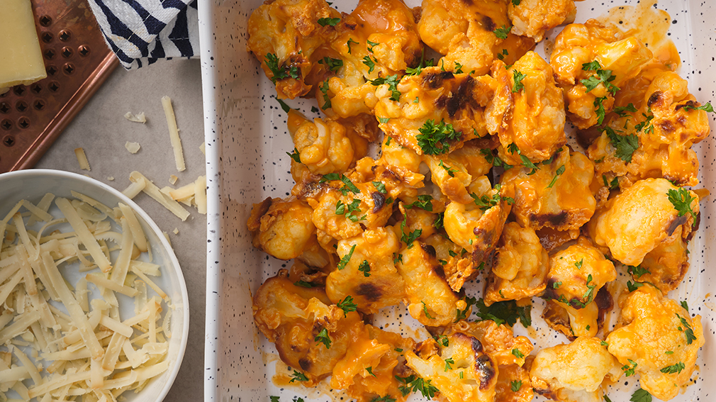 Cheese crusted cauliflower wings in white speckled pan next to white bowl of shredded white cheese.