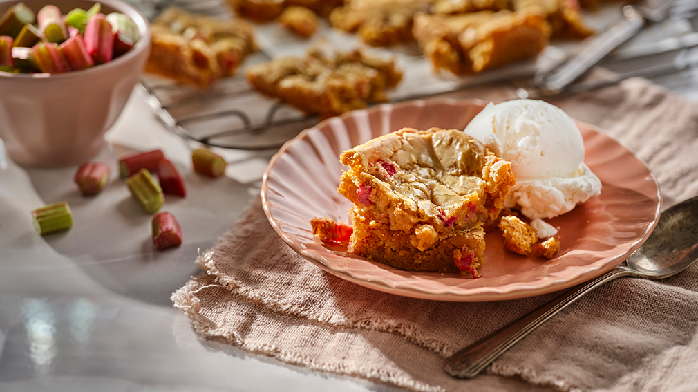 A square of an Ultimate Rhubarb and White Chocolate Blondies sits on a peach coloured plate, next to a scoop of ice cream.