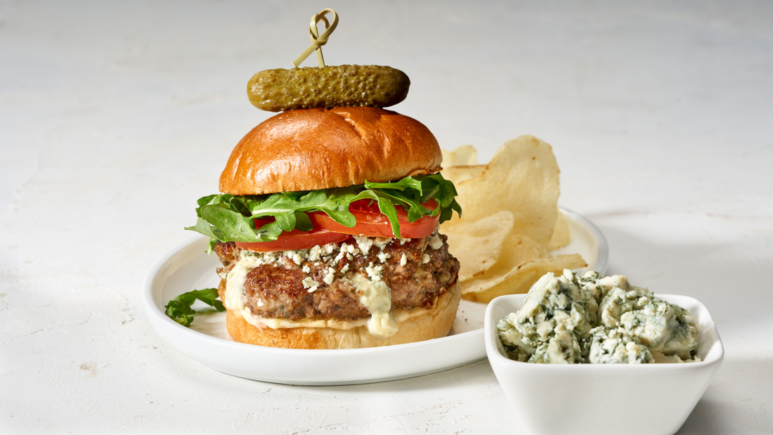 A small white plate with a tall Blue Cheese-Stuffed Burger with a gherkin pickle placed on top of the burger and a stick poking through the pickle and the burger. Also on the plate is a handful of chi