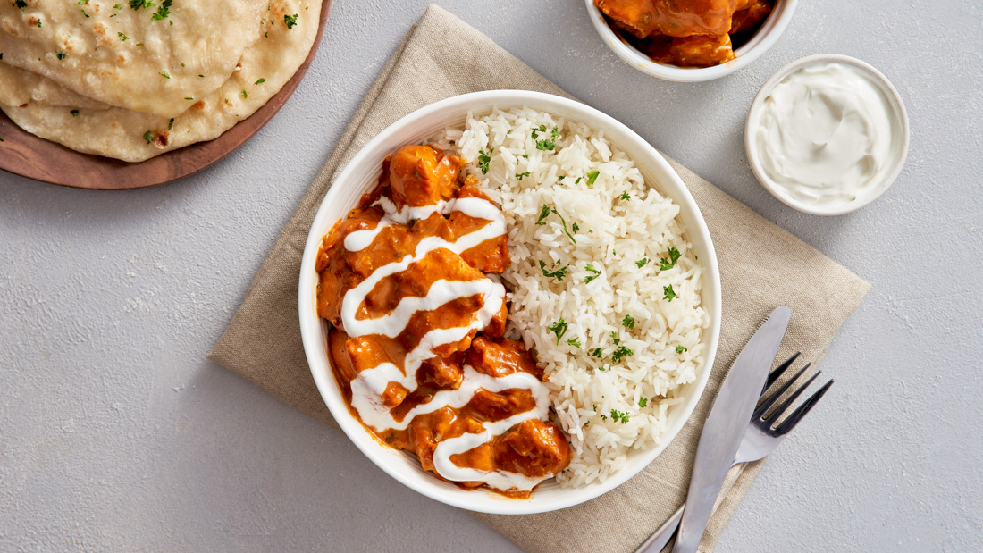 White plate with vibrant Butter Chicken drizzled with plain yogurt covering the left side of the plate and white rice garnished with coriander covering the right side of the plate.