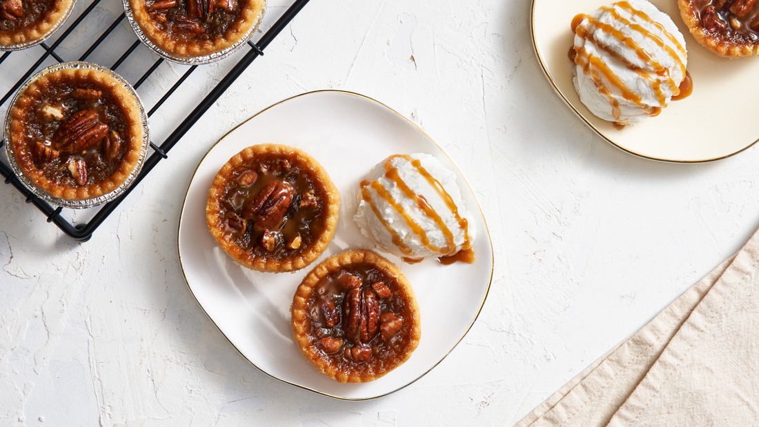 Two white plates each with two Caramel Pecan Butter Tarts and a scoop of Ontario Vanilla Ice Cream and all drizzled with caramel sauce.