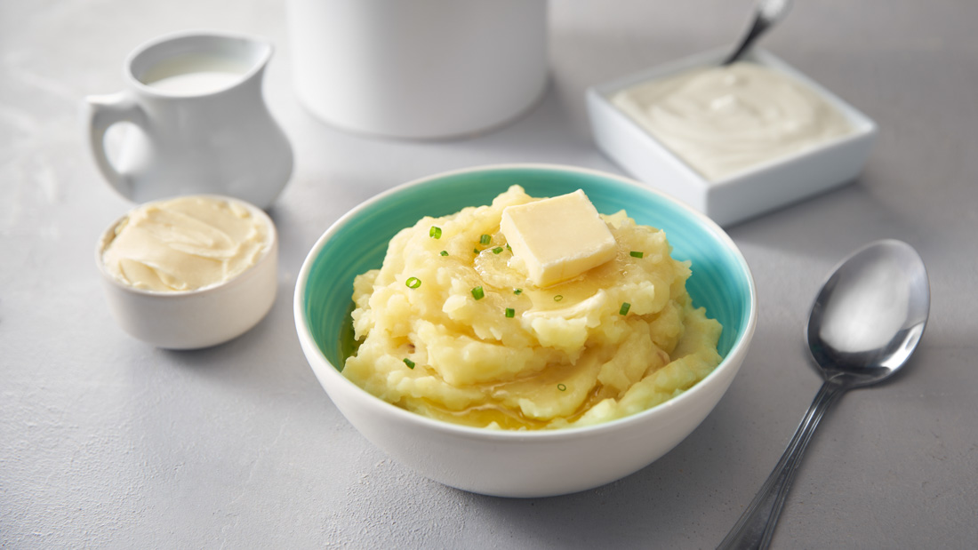 A white and aqua blue bowl filled with Mashed Potatoes, topped with a small slice of butter and garnished with chives. Sour cream in a small bowl on the side beside the bowl of mashed potatoes.
