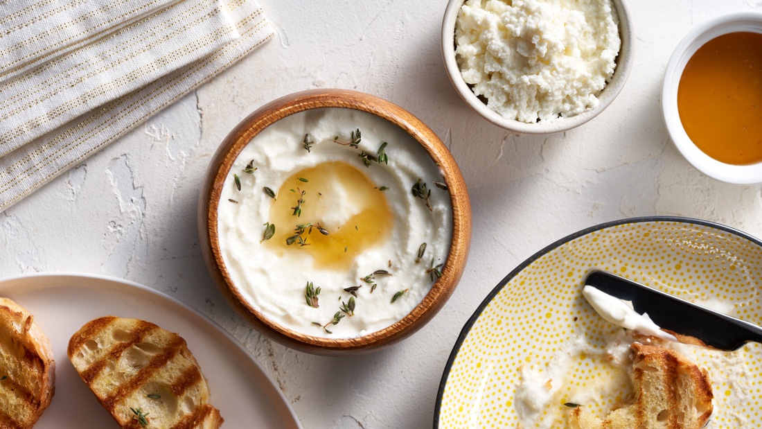 A small wooden bowl filled with Whipped Ricotta with a dollop of Honey in the middle and garnished with thyme leaves on a lightly textured white countertop.