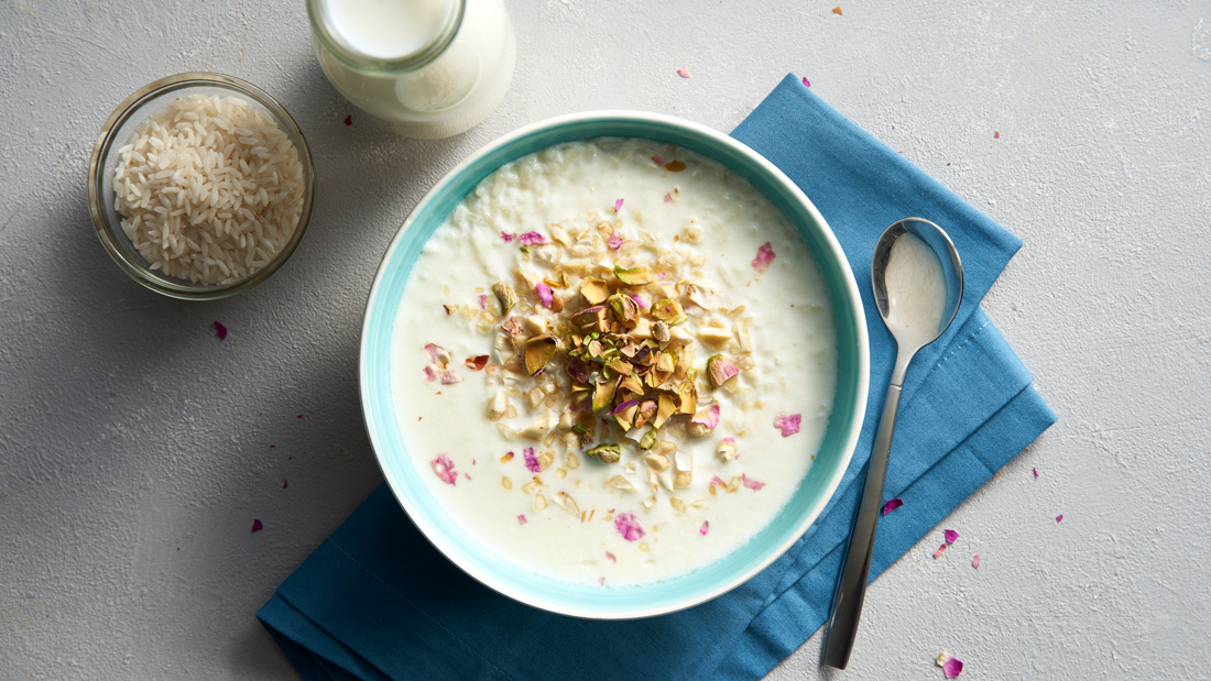 A light blue bowl filled with Kheer Rice Pudding and topped with chopped nuts and rose petals on a white countertop with a long silver spoon.