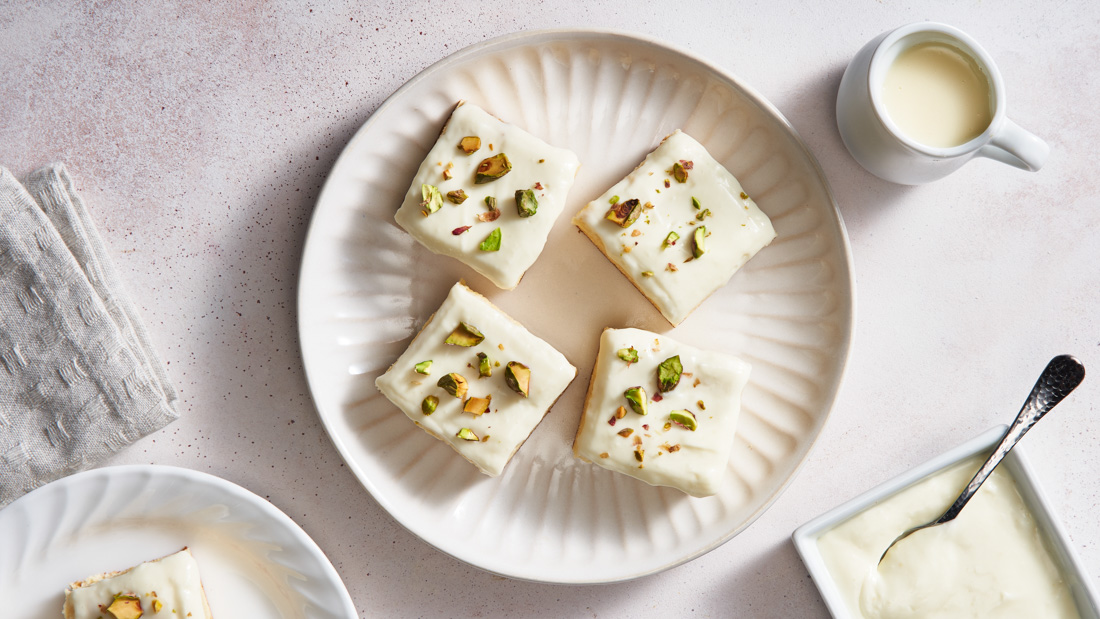 A white plate on a white tabletop with four Middle Eastern Milk Cake Slices garnished and topped with crushed pistachios.