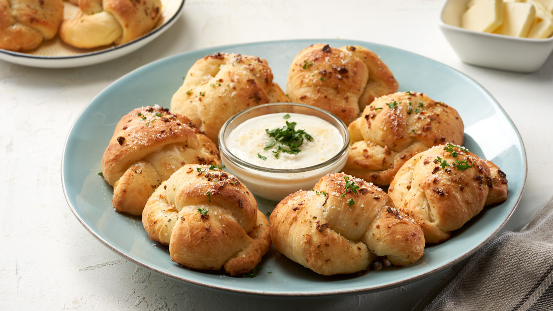 A light blue plate filled with seven parmesan garlic knots brushed with garlic butter and garnished with fresh parsley and Ontario Parmesan all organized in a circle around a small clear glass bowl of